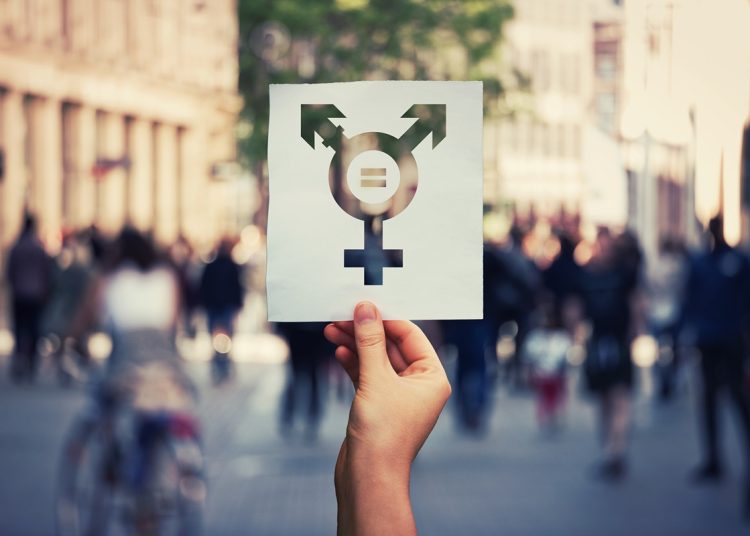 Hand holding a paper sheet with transgender symbol and equal sign inside. Equality between genders concept over a crowded city street background. Sex rights as a metaphor of social issue.