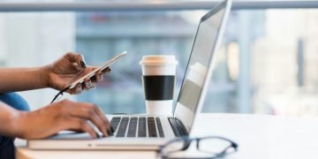 hands typing on laptop while holding mobile phone
