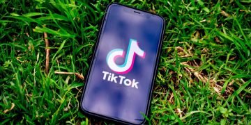 A mobile phone lies on the grass with its screen set to the TikTok home page