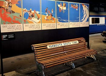 A wooden bench labelled Yarmouth South Town sits beneath a panel of six posters that make up one image of people enjoying the coast