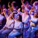 older people sitting on chairs holding their right arms above their heads as if to shield themselves