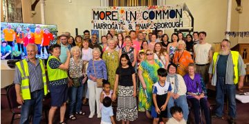 Large diverse group of people of all ages posing for a group photo in a hall, with a colorful 'More In Common' banner reading Harrogate & Knaresborough behind them.