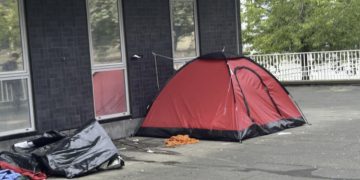 Red dome tent pitched on a concrete sidewalk beside a dark brick building, with scattered tarps and clothing nearby - this is the site of a rough sleepers’ encampment in Bradford 2026