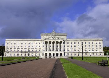 Front view of a grand white neoclassical government building with a colonnaded entrance and steps, set in green lawns and pathways under a cloudy sky.