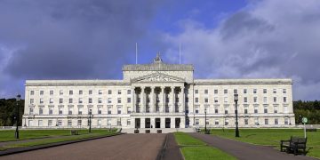 Front view of a grand white neoclassical government building with a colonnaded entrance and steps, set in green lawns and pathways under a cloudy sky.
