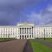 Front view of a grand white neoclassical government building with a colonnaded entrance and steps, set in green lawns and pathways under a cloudy sky.
